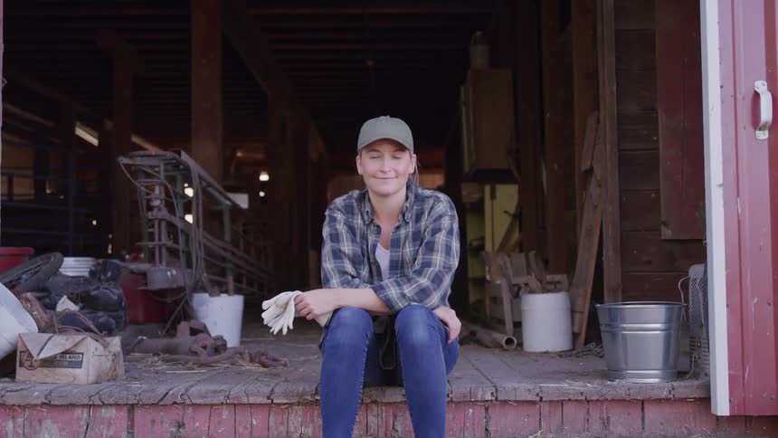 A woman in a plaid shirt and cap sits on the steps of a barn smiling calmly