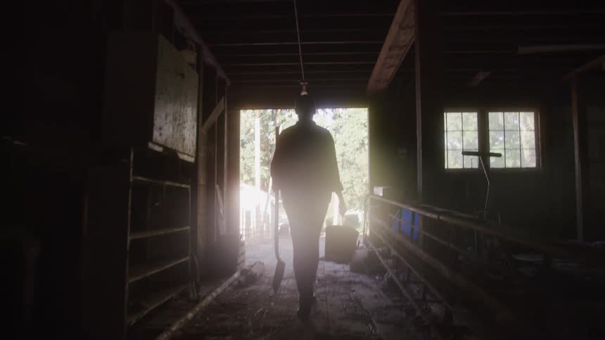 A person walks away from the camera through a dimly lit barn or shed carrying a shovel and a bucket towards a bright outdoor light