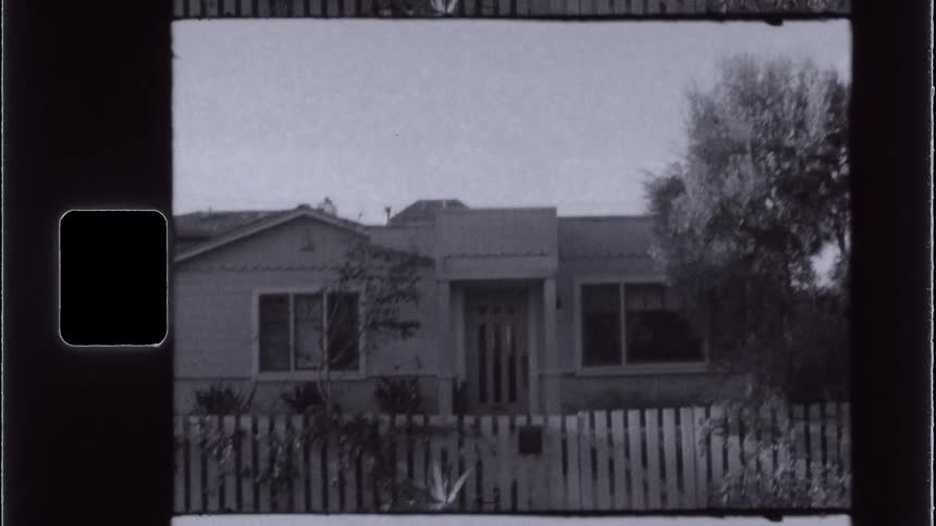 A blackandwhite film frame shows a small house with a white picket fence