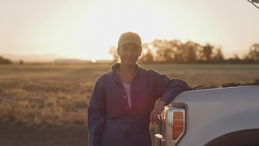 A woman standing leaning against a car with a gentle smile under sunlight