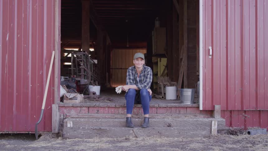 A young woman sits at the entrance of a barn wearing a hat jeans and holding work gloves