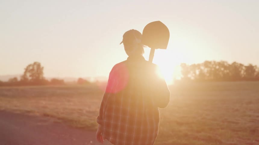 Woman in cap walking with a shovel on her shoulder in an agricultural setting at dawn