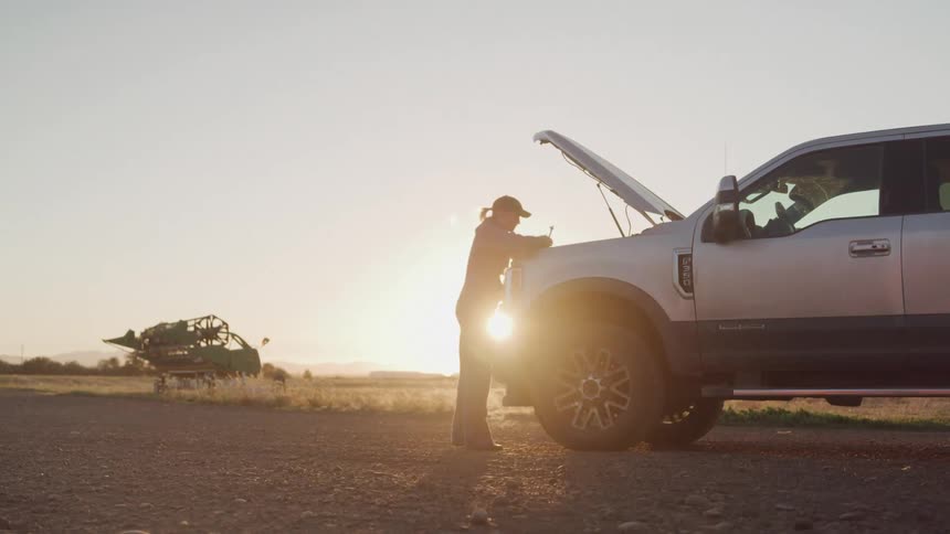 A young woman wearing a baseball hat checks the engine of her car on the side of the road on a sunny day