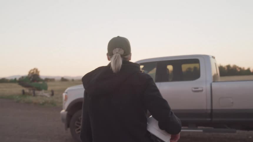 Woman carrying a white helmet in her hand and walking towards a pickup truck in a field