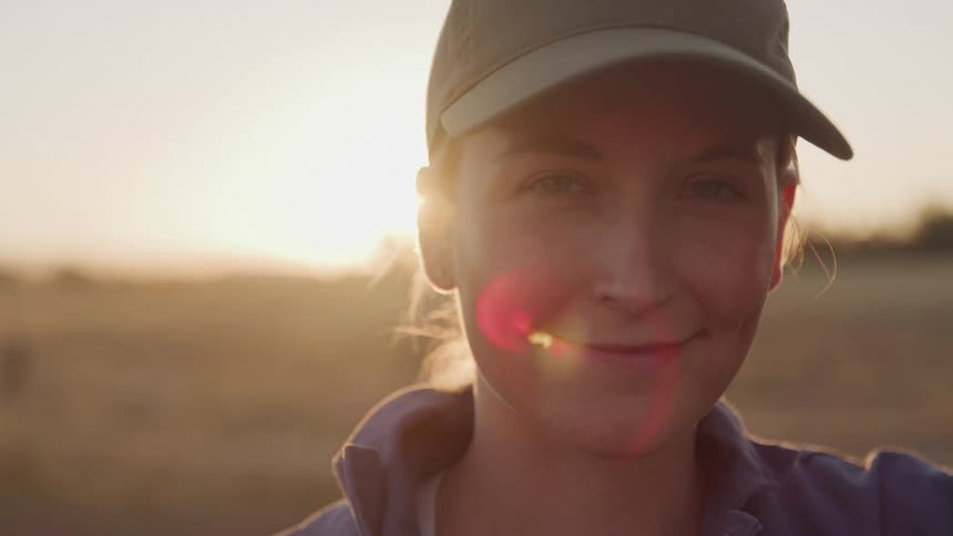 A woman in a cap smiles softly at sunset in an open field