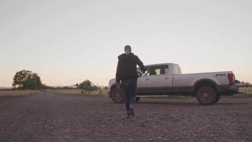 A woman walks down a gravel road toward a silver ford f150 parked in a rural landscape bathed in sunset light