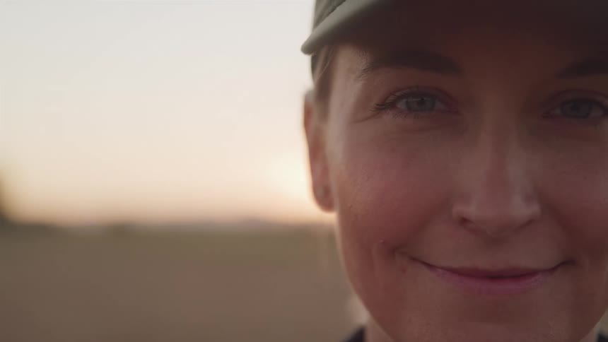 A woman wearing a hat stands outdoors at sunset against a pink sky