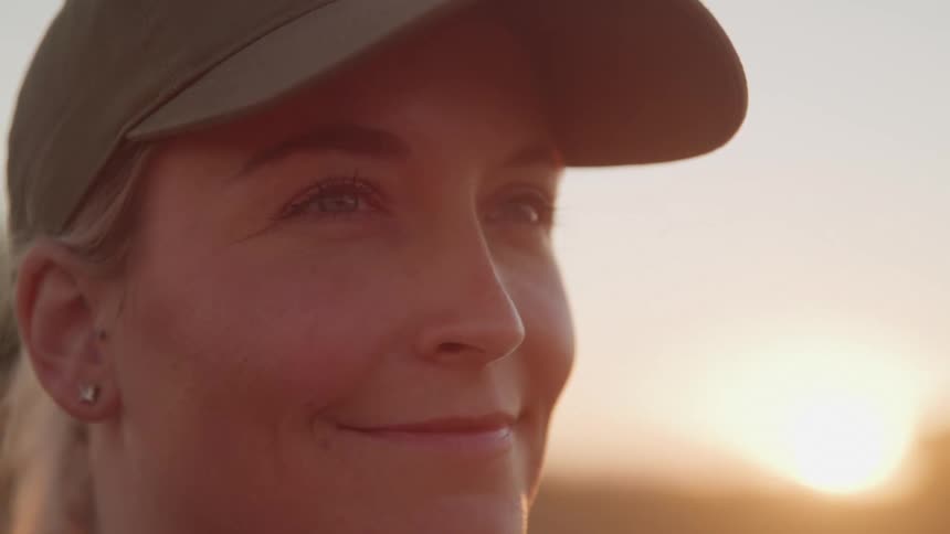 A woman wearing a dark cap looks to her right with a gentle smile with the warm glow of sunset in the background