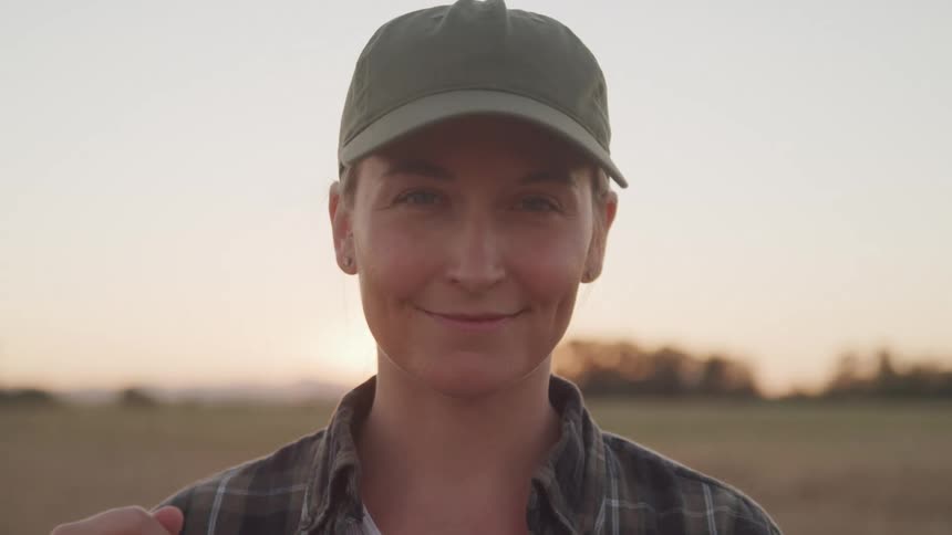 A smiling woman in a cap stands in a field at sunset