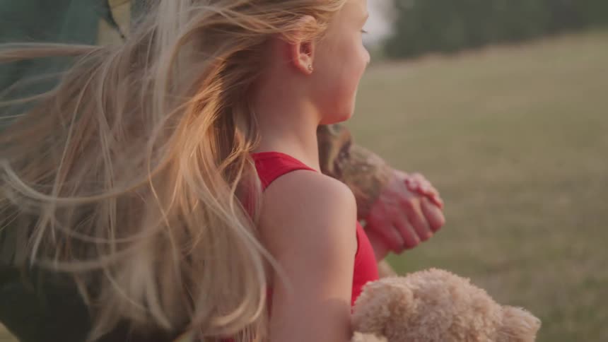 Little girl accompanied by an adult walking in a field and carrying a small bear