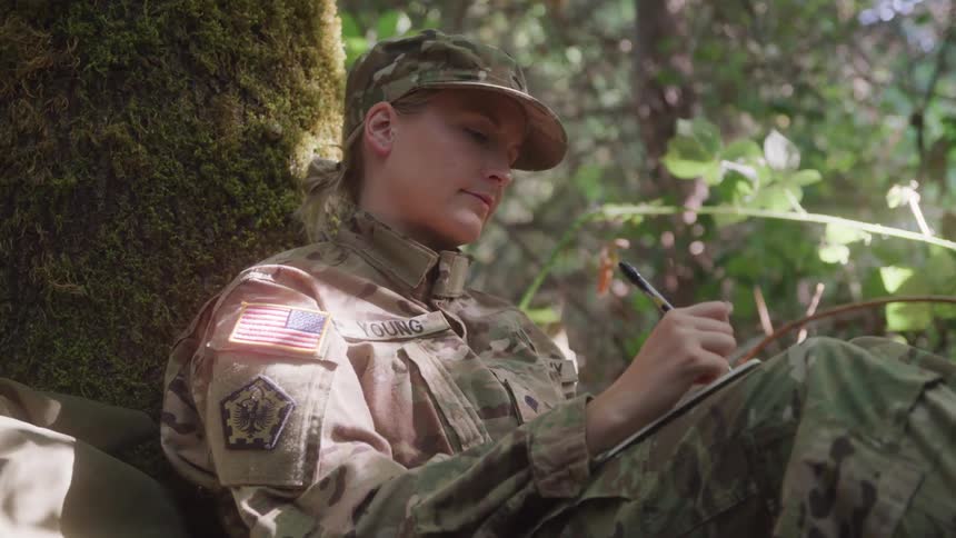 A us military servicewoman sits in the forest writing a letter by hand