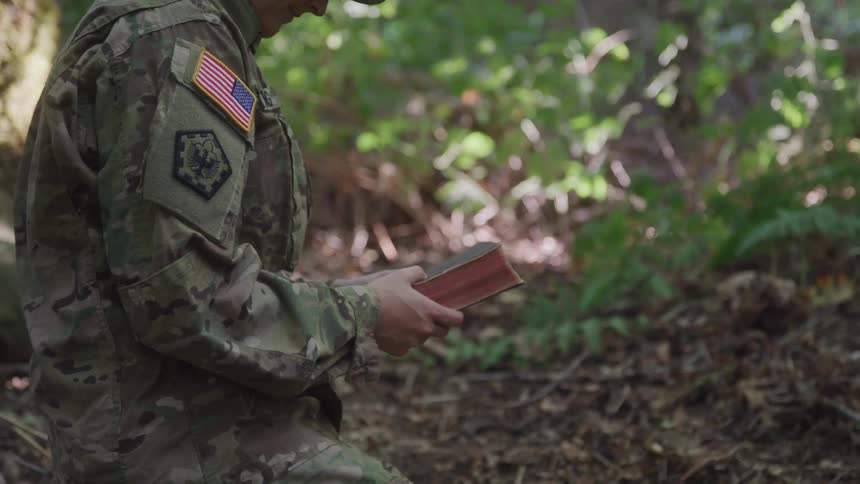 A soldier is kneeling in the dirt while holding a book
