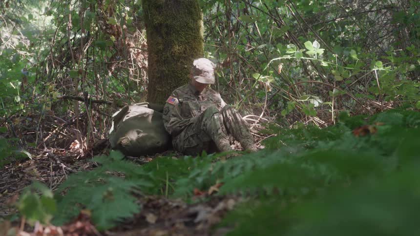 A soldier sits alone in a forest reading or writing near a tree