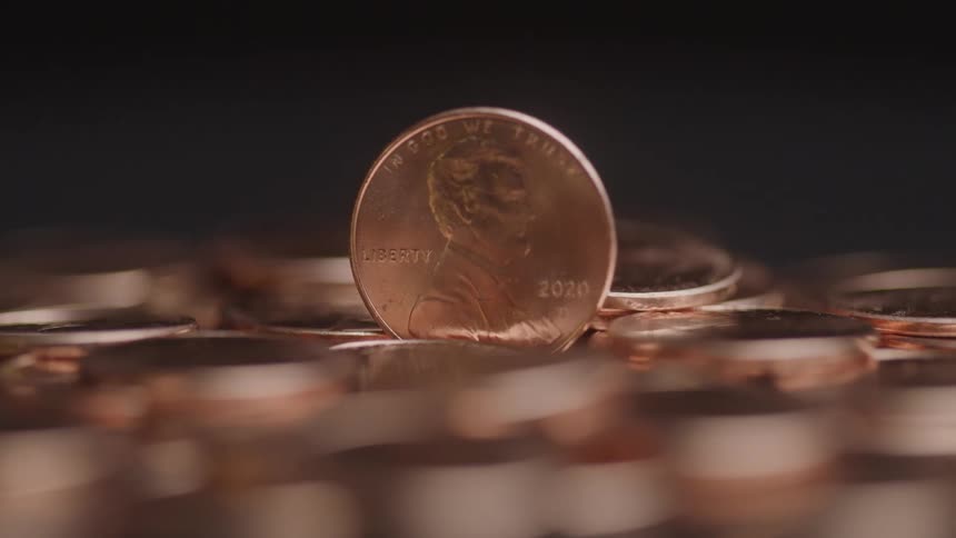 A closeup of a single penny stands upright among a pile of scattered pennies