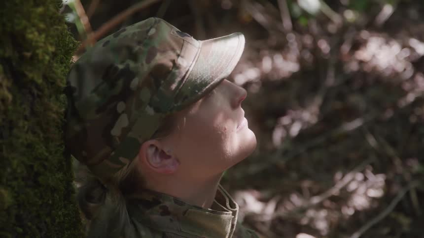Woman in military uniform sitting against a tree in the forest with the sun reflecting off of her face