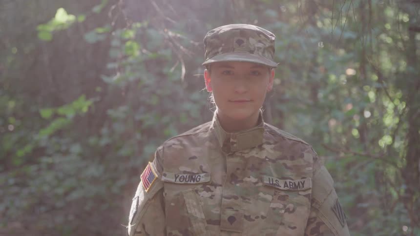 A army soldier stands in uniform in a sunlit forest looking calmly at the camera