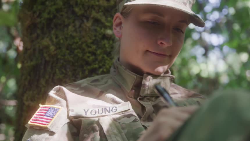 A young female soldier in camouflage uniform and cap smiles as she writes in a notebook while resting against a tree in the woods