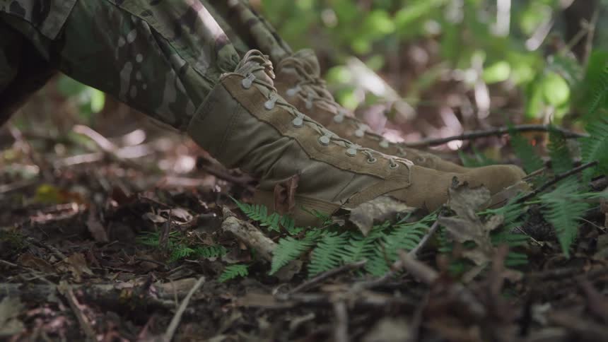 A person wearing a military uniform and boots sits on the forest floor around midday