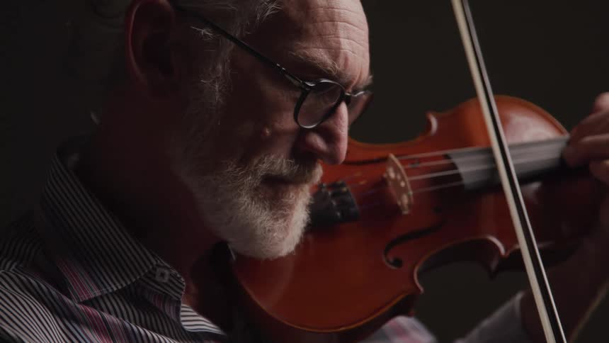 An elderly man plays the violin with focused emotion in a dimly lit setting