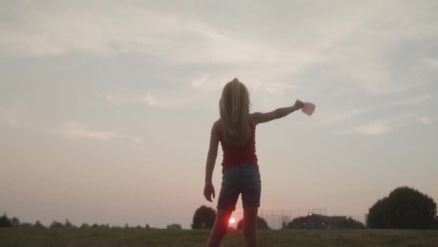 A little girl waves a mini american flag in an empty field at sunrise