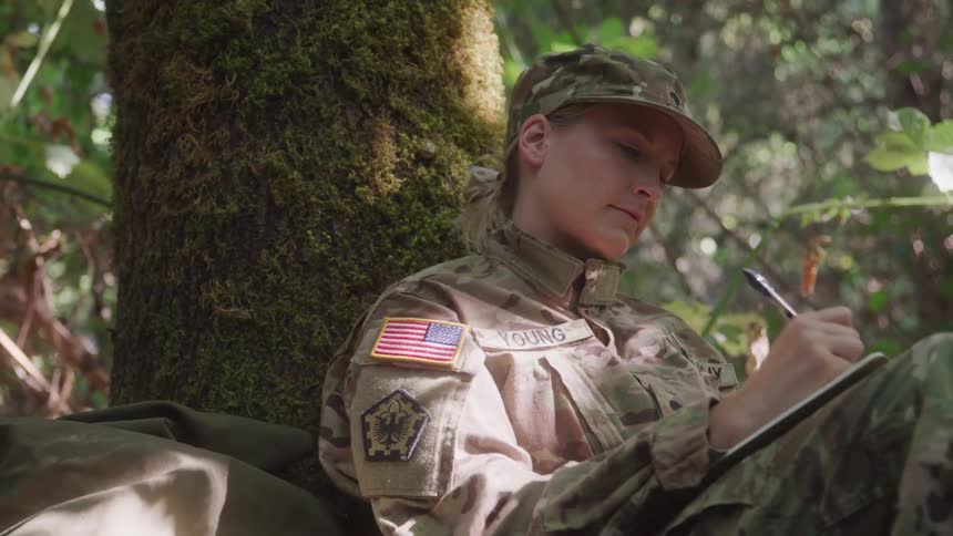 Military woman writes in a notebook while resting against a tree in the forest
