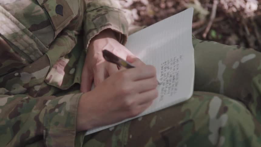 A person in camouflage uniform sits outdoors writing in a notebook