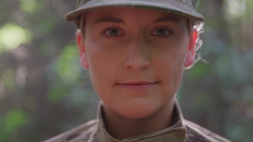A female soldier in uniform looks calmly into the camera in a sunlit forest