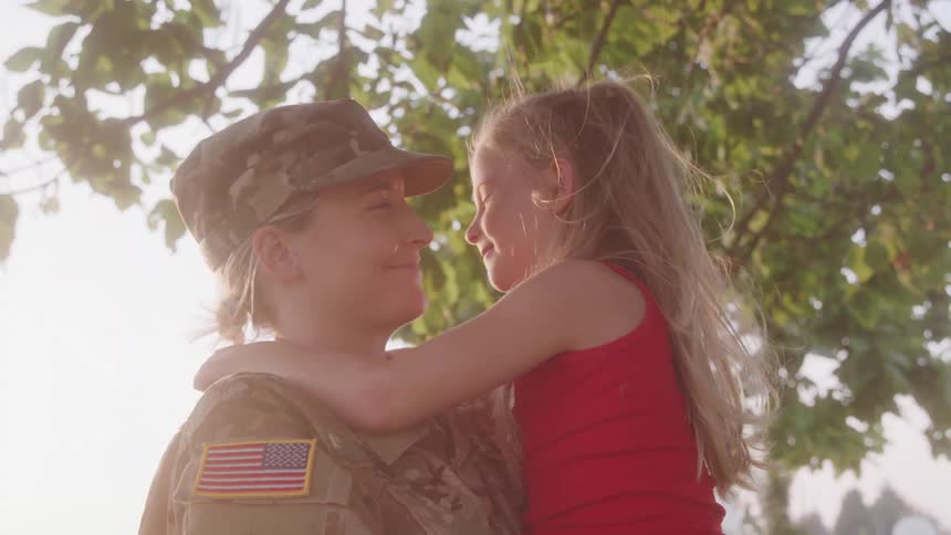 A woman in military uniform with an american flag patch holds a child in a red sleeveless shirt against a backdrop of dense green foliage