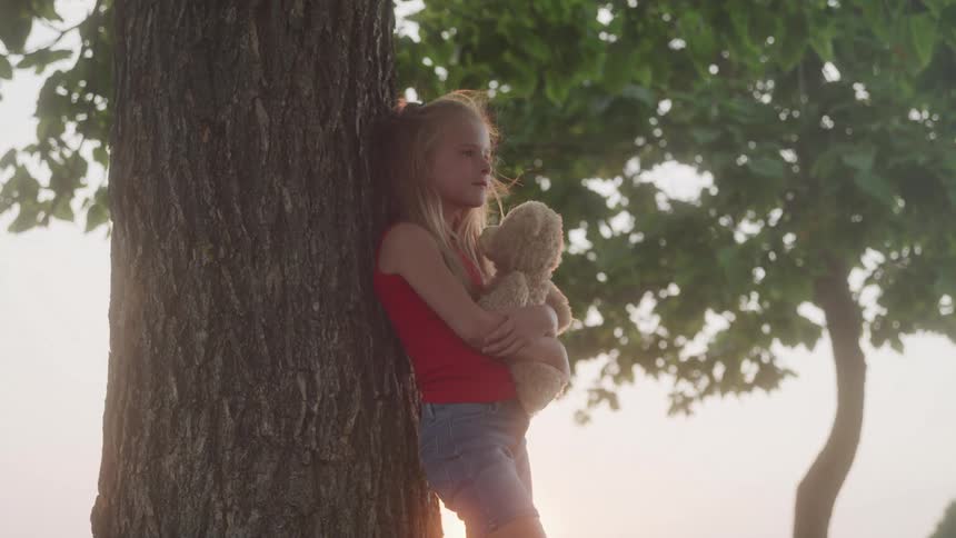 Young girl leans on tree holding teddy bear looking thoughtful