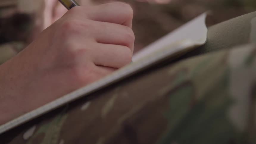 A soldiers hand grips a pen while writing in a small notebook resting on camouflaged pant legs