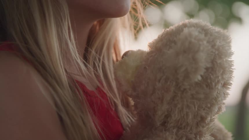A girl in a red shirt holds a beige teddy bear close to her chest with blurred green foliage in the background
