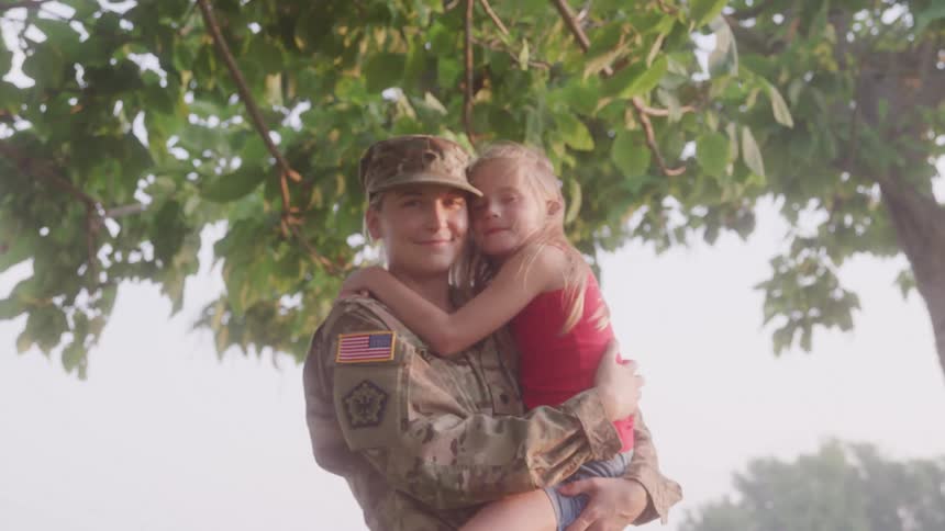 A soldier in uniform holds and hugs a smiling young girl under a tree