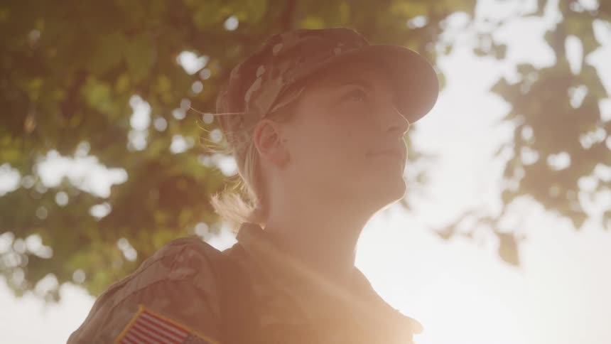 A woman in military fatigues and a camouflage cap looks upwards with a serene expression with tree leaves and a bright sky in the background