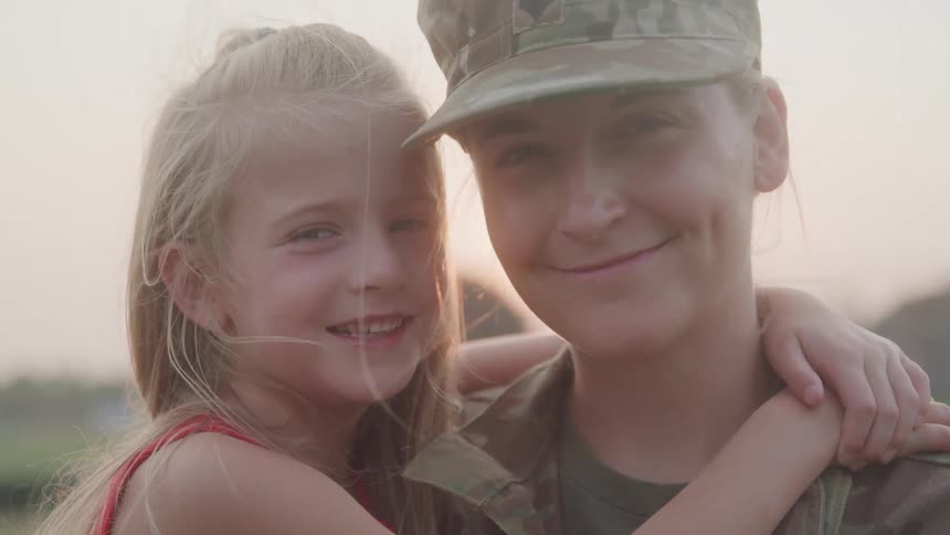A female soldier in uniform hugs a smiling young girl in a warm outdoor setting