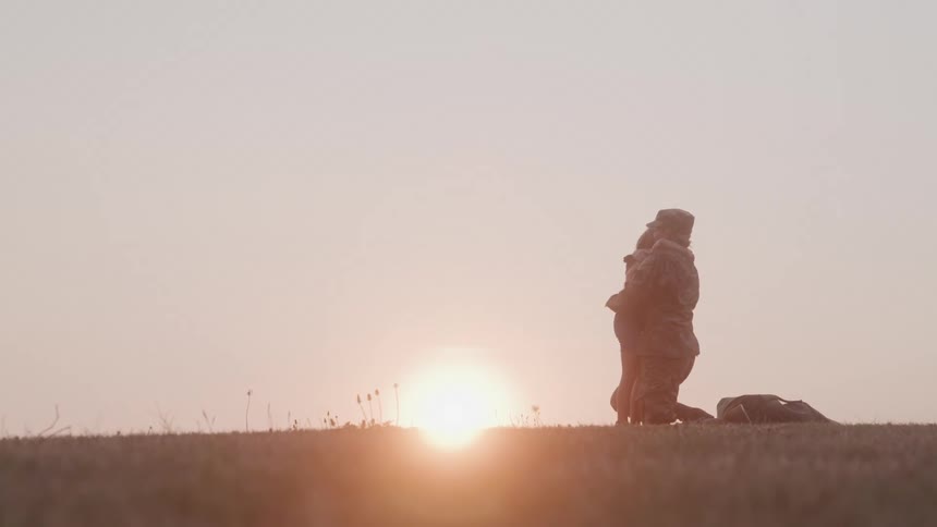 A military mother drops her bag on the grass and kneels down with her arms open to hug her daughter who runs to her they embrace with happiness as the sun sets in the background