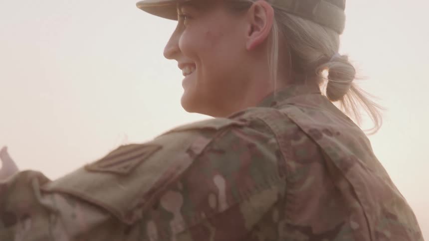 A person in military uniform holds a child in a red shirt in the glow of a sunset