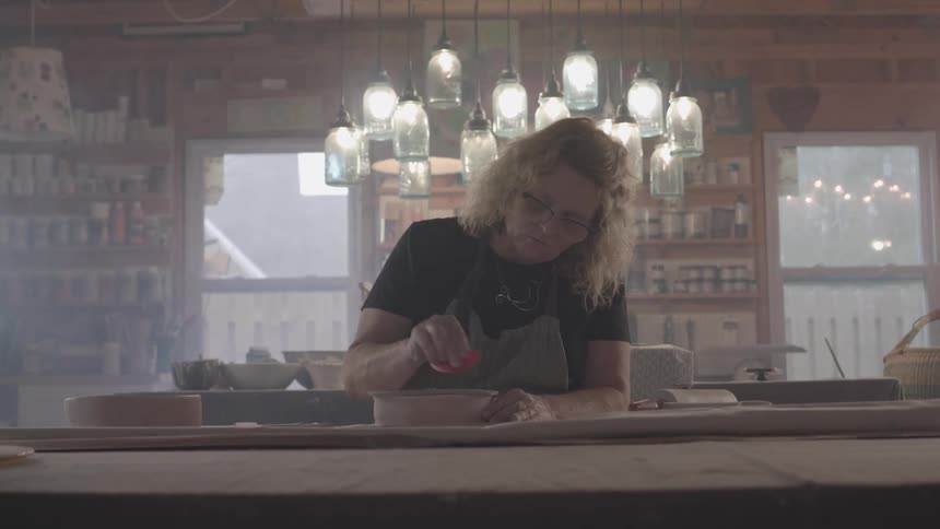 A woman shapes clay by hand in a cozy pottery studio under warm lighting