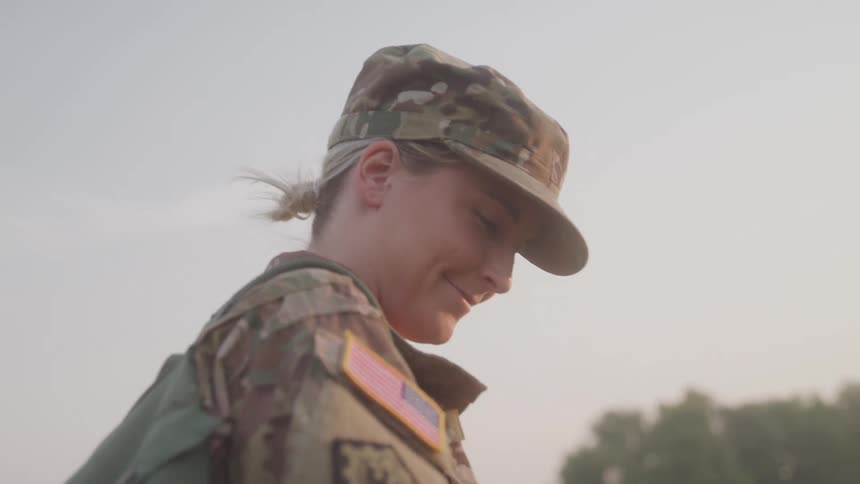 A military woman wearing a uniform and a hat walks under the sun with a smile around midday