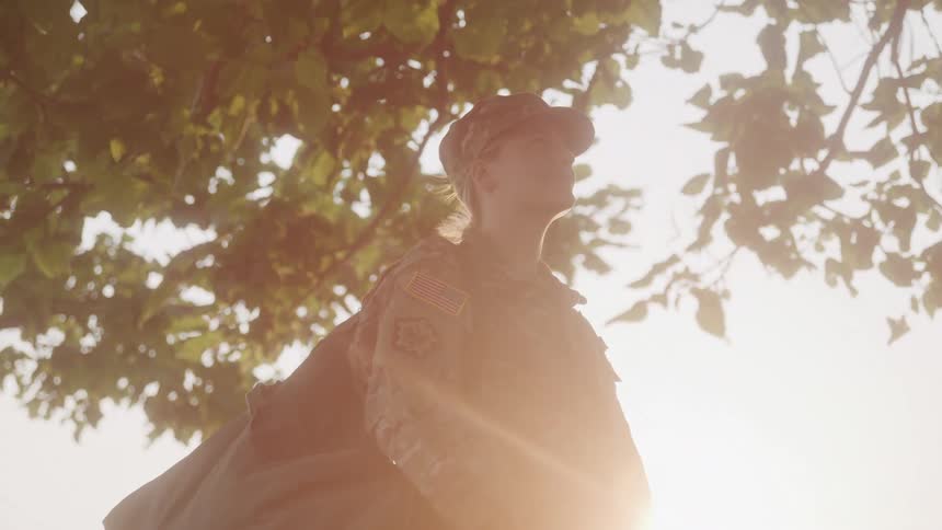 A woman in military uniform with a camouflage cap looks upward with green leaves and a bright sky in the background