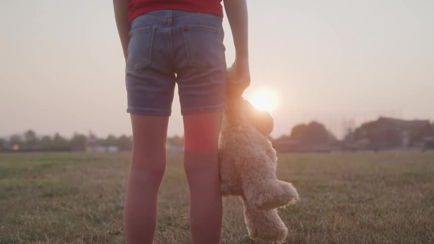 A child stands in a field at sunset holding a teddy bear