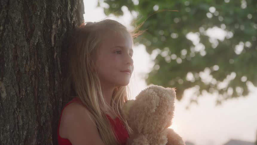 A young girl with long blonde hair leans against a tree trunk holding a teddy bear and looks towards the right with a soft expression