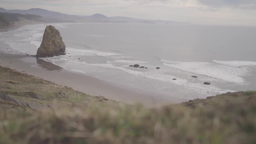 A large rock formation stands along a quiet coastal beach with waves gently washing ashore