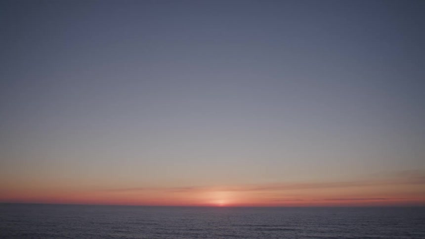The sun partially appears on the horizon over the sea illuminating the sky with soft hues of orange pink and blue