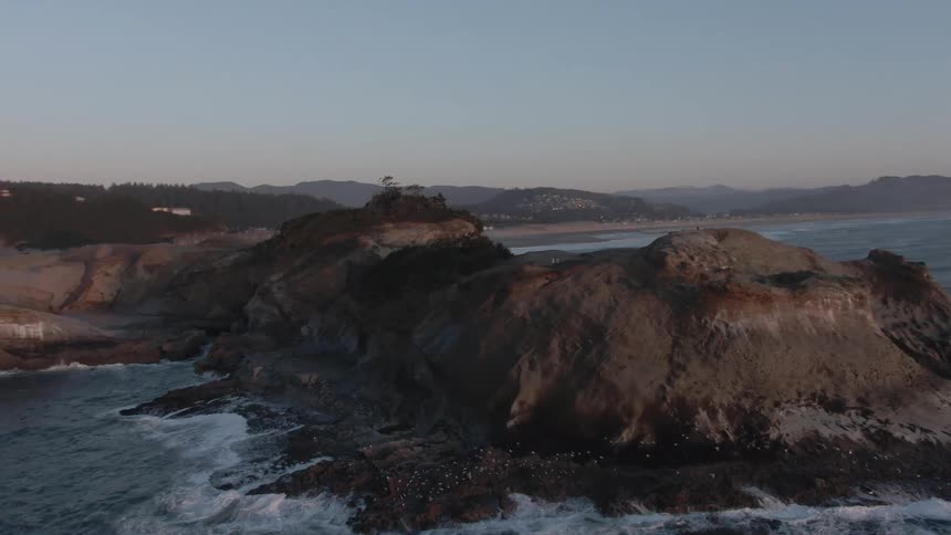 Rocky coastal cliffs meet the ocean at dusk backed by forested hills and distant homes