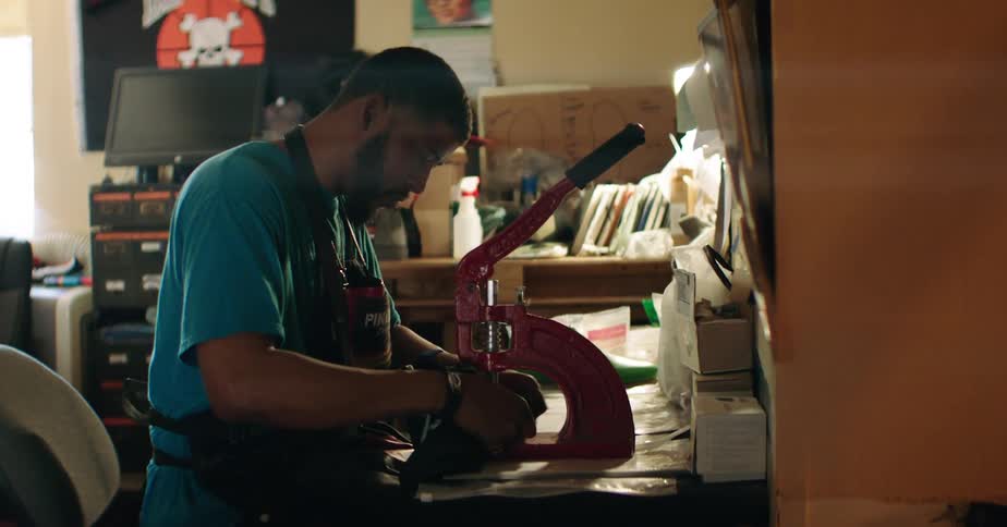 Man operates a hand press at a workbench surrounded by tools boxes and sewing supplies