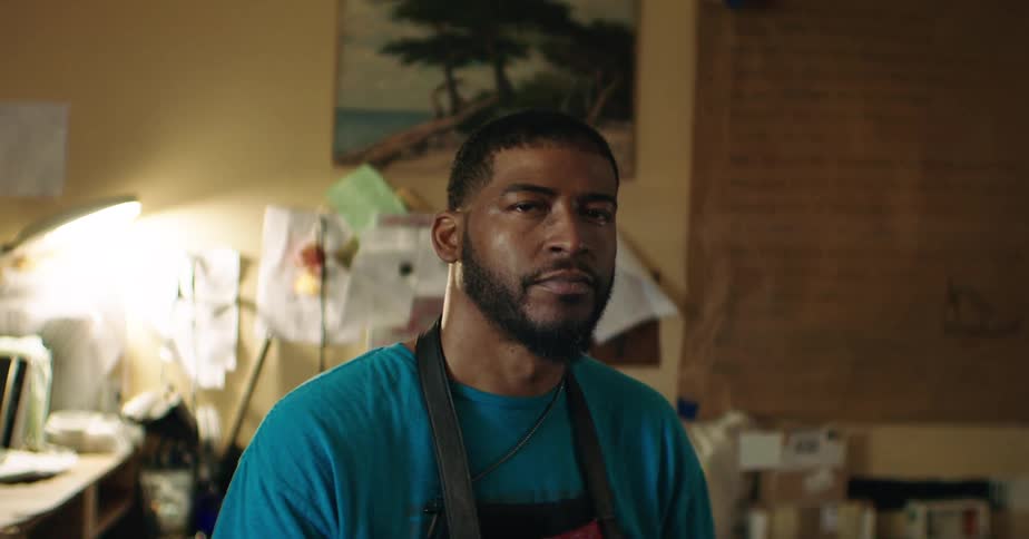 A man wearing an apron sits in a warmly lit workspace looking directly at the camera
