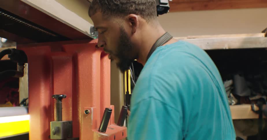 A man wearing a blue shirt and work gloves operates a press in a cluttered workshop