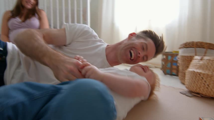 A father lies on the floor tickling his young son while a pregnant mother sits nearby in a softly lit nursery with a white crib