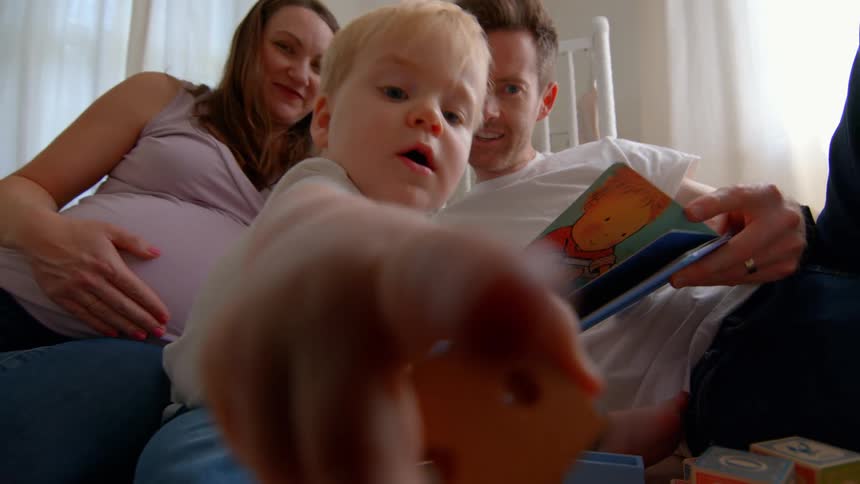 A family sits on the nursery floor reading together while the toddler plays with toys