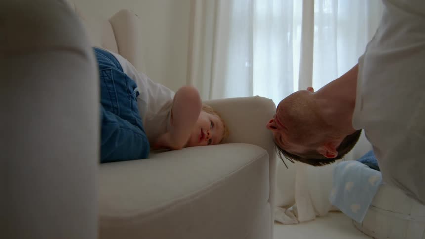 A father leans toward his son playfully as the child relaxes on a sofa in a softly lit living room